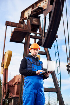 Vertical portrait of oil man, wearing blue overalls and orange helmet, with a laptop, standing with his back to an oil rig, checking oil pumping unit, making notes in his computer