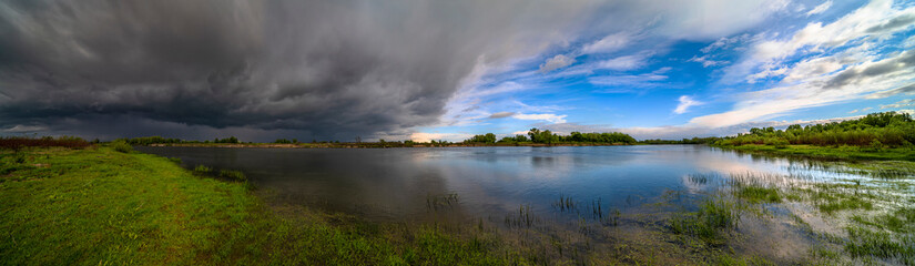 Naklejka na ściany i meble Amazing landscape of nature before thunder storm. Dark clouds cover blue sky at wild river. Incredible weather panorama
