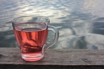 Glass cup of herbal tea with a pink tea rose petals on the wooden pier over the river. Summer tea party outdoors