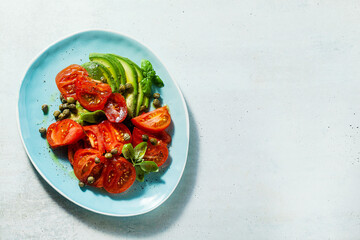 summer salad with ripe tomatoes and avocado with capers in a blue plate on the table