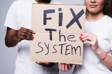 Young african man and coucasian woman holding a cardboard poster with the message text Fix the system isolated on white background. Concept on the theme of protest for police brutality and racism.