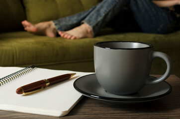 Gray cup of coffee, blank notebook and pen on the desk, woman relaxing on the green sofa.Concept of work and rest