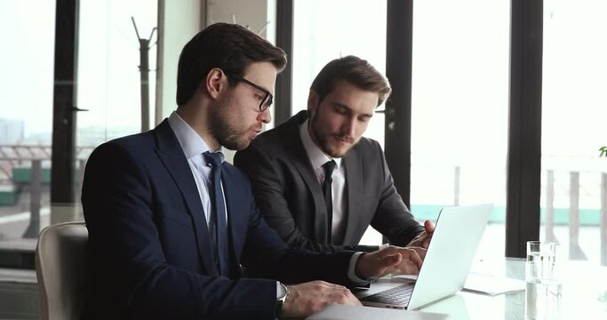Two serious businessmen partners wear suits discuss online ebusiness startup idea using laptop looking at computer screen. Male executives, manager and client work together analyze financial market.