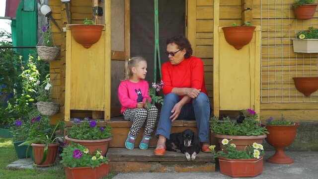 Cute Little Girl Talking with her Grandmother while Sitting on the Steps of Country House. Concept of Family, Generations and Happy Childhood