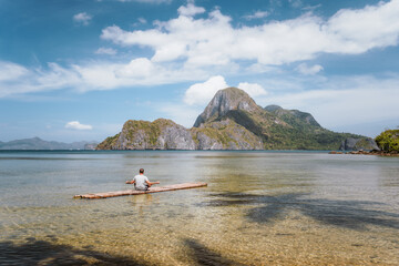 Fototapeta na wymiar Man meditating on bamboo float in shallow lagoon water and Islands of Cadlao bay in a distance. Palawan, Philippines. Holiday vacation concept