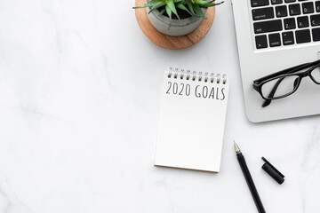 Small blank notebook with 2020 goals text is on top of white marble office desk table with office supplies. New year plan and resolutions concept.