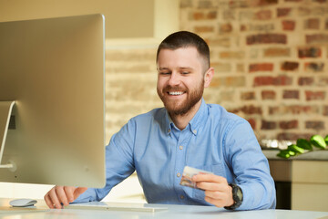 A man with a beard in a blue shirt smiles looking at his credit card in his hands at home. A guy doing an online payment on the internet on a desktop computer in his apartment. 