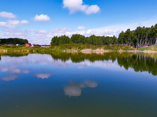 A small pond in the village. Reflection in the water of a blue sky with clouds. Hot summer sunny day. Picturesque traveling, seasonal and nature beauty concept scene.