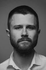 A close portrait of a bearded brutal man in a white shirt who is standing in front of a grey background. A serious guy is looking right into a frame. A black and white photo.