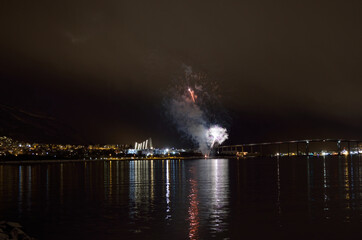 Naklejka na ściany i meble Beautiful firework on night sky in tromsoe city with bridge, cathedral and colorful reflection on the cold fjord water surface