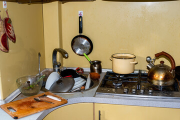 A scruffy and dirty kitchen, a stove in grease, in the sink a mountain of unwashed dishes. Unsanitary conditions, a mess in the house. The kettle and cutlets are all burnt and plaque, plates spotted