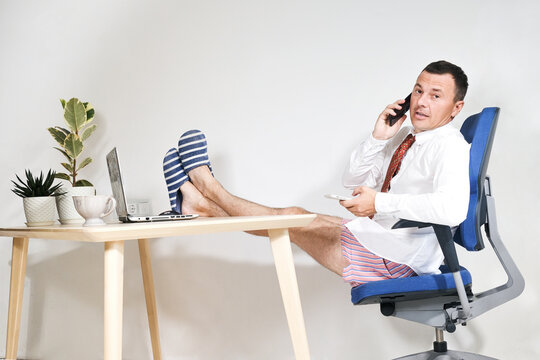 A Male Manager Works At Home Remotely, Holds An Online Meeting. He Is Wearing A Shirt And Tie And Homemade Shorts And Slippers. Stay Home And Quarantine. Remote Work, Work On The Internet, Remotely