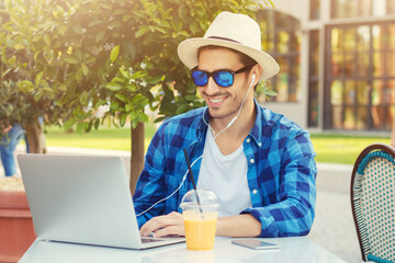 Outdoor portrait of young smiling european man sitting at cafe table, his laptop open, listening to music through earphones, drinking juice and enjoying summer time outside