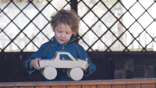 A small boy in a blue denim shirt plays with a homemade cardboard car in a wooden gazebo. Child in the open air.
