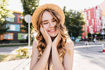 Cute girl in hat laughing with eyes closed on city background. Romantic white woman dreamy posing in summer morning.