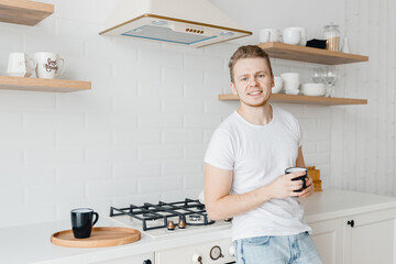 Portrait young man smiles drink fresh coffee in ceramic black mug breakfast. Bright kitchen background