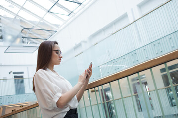 A young positive business woman in glasses and a white shirt uses a smartphone to communicate with colleagues,