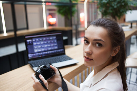 young attractive girl is sitting in front of a laptop and holding a camera in her hands. Photographer and Retoucher