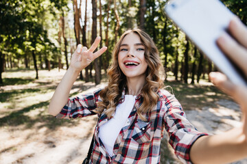 Glamorous curly girl posing with smile in forest. Charming female model using smartphone for selfie on nature background.