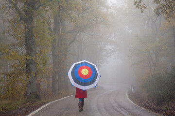 Girl with a red coat and an archery shooting target umbrella walking on an empty road surrounded by...