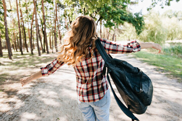 Portrait from back of debonair woman with leather backpack. Outdoor photo of amazing curly girl in checkered shirt dancing in park.