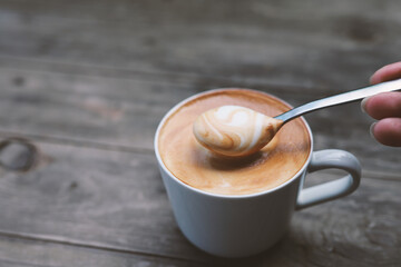 Hand holding a spoon that scoop the coffee in a cup. Cup of Cappuccino coffee with a delicious crema.