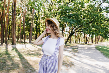 Happy female model in cute summer attire posing in park. Outdoor shot of shy caucasian girl in hat.