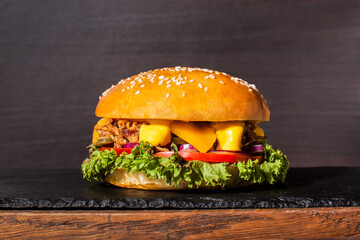 Burger with pork cutlet lies on a board with a stone. And pickles, cheese, red onion, mustard and fresh salad leaf. Dark wooden background at the back