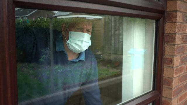 Old Man Wearing Face Mask, PPE. Looks Out Of Nursing, Care Home Window.  High Risk, Self Isolation And Protection From Coronavirus, Covid-19. 