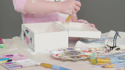 Little girl painting a white unicorn with acrylic paint on a wooden box.