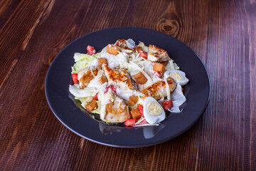 Delicious and tender salad with chicken and fresh vegetables dressed with sauce in a black plate on wooden table background. Warm salad from chicken, tomato and quail egg. Dietary menu
