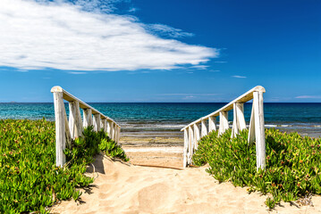 Entrace to the deserted sandy beach with wooden railing in green grass by sides. At the background green blue sea and blue sky with light white clounds. Sunny summer day.