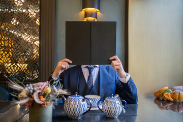 Man sitting in restaurant in suit holds book on level heads. A man in a suit holding a plate instead of his head.