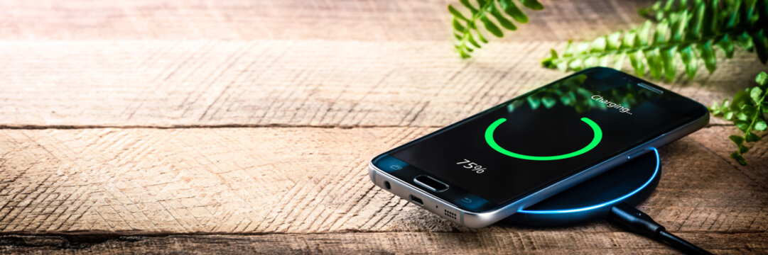 Smartphone On Wireless Charging Station On Wooden Desk With Green Plant