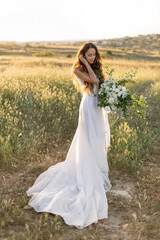 Fototapeta na wymiar Refined slender bride in luxury fashion wedding dress holds a large wedding bouquet of peonies and wild flowers. Charming bride in a flowing dress stands in a green field of spikelets in the sunlight.