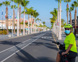 Woman with medical mask riding her bicycle in the deserted road in Tenerife, Canary islands. No tourists because of the Covid-19 coronavirus. Palm trees and blue sky in background