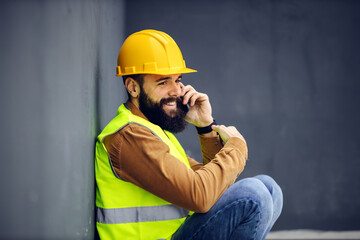 Young smiling friendly bearded worker in vest, with helmet on head crouching and leaning on the wall while having a phone conversation on lunch break.