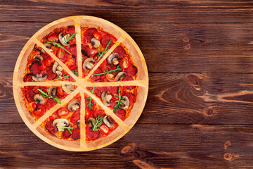 Fototapeta na wymiar Sliced hot pizza with salami, arugula, cherry tomatoes, mushrooms and texas spice mix, on a round wood platter which is on wooden rustic background, top view and copy space