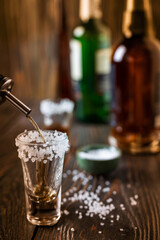An alcoholic drink at the bar counter is poured into a tequila glass with salt at the edges, in the background are different bottles of alcohol, shallow depth of field, selective focus. The concept of
