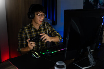 Fototapeta na wymiar Handsome excited asian gamer guy in headphones enjoy and rejoicing while playing video games on computer in cozy room is lit with warm and neon light, gaming and technology e-sport concept
