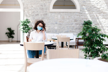 Social distancing during quarantine. Young woman wearing protective mask in cafe indoors. Girl talking on mobile phone. Lifestyle moment. Restaurant with safety space between seats in new normal.