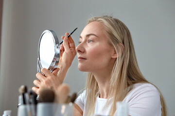 Blue-eyed blonde does make-up at home. Draw your eyebrows with a brush.