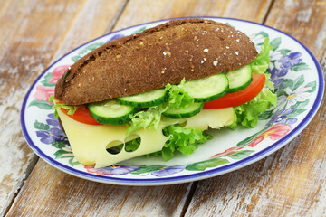 Whole grain roll with Swiss cheese, lettuce, tomato and cucumber on colorful vintage plate on wooden surface
