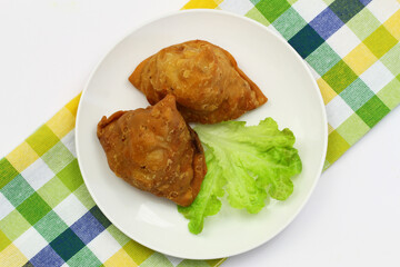 Two Indian samosas on green lettuce on white plate on checkered cloth
