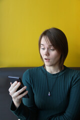 A young beautiful girl of European appearance in a blue jacket sits in a cafe with a mobile phone on the couch against the background of a yellow wall. Surprised expression. Place for text, copy space