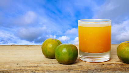 a glass of orange juice and a few oranges are on the wooden table in the blue sky with clounds background