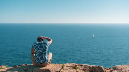Young man with his hand on his head sitting on a cliff with the sea in front of him.