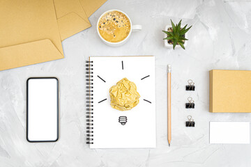 Refreshment break idea and productivity boost concept. Flat lay of an office workplace with a smartphone, coffee cup, light bulb symbol made of a crumpled paper ball and a sketch on a spiral notebook.