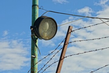 Obraz premium one old round spotlight with a lamp on an iron pole at the fence with barbed wire against the sky and clouds