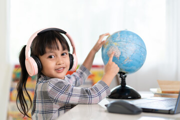 Asian beautiful little student girl is smiling and pointing on bilingual globe model for her online lesson, concept of learn from home and homeschooling for child education. - obrazy, fototapety, plakaty
